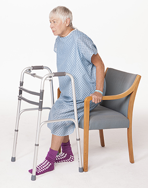 Mujer vestida con una bata de hospital sentándose en una silla con un andador al frente.