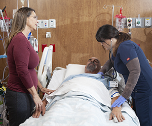 Proveedor de atención médica atendiendo a un hombre en una cama de la unidad de cuidados intensivos mientras una mujer está de pie cerca.
