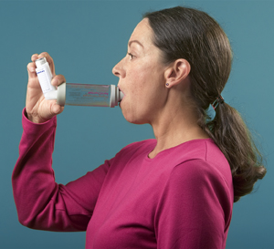 Side view of woman with spacer in mouth. Inhaler is inserted into other end of spacer. She is pressing on end of inhaler.