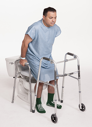 Man in hospital gown in bathroom holding onto toilet side rails to help steady him while he sits on toilet.