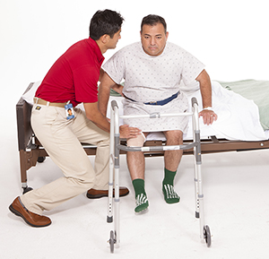 Proveedor de atención médica que ayuda a un hombre a ponerse de pie desde una cama de hospital; hay una andadera cerca.