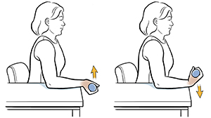 Una mujer sentada en una silla con el brazo apoyado sobre la mesa hace un ejercicio de flexión de la muñeca con una mancuerna.