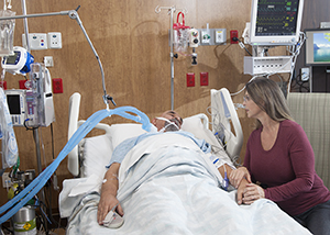 Mujer sentada junto a la cama de un hombre intubado en una unidad de cuidados intensivos.