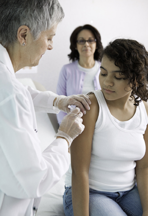 Proveedor de atención médica aplicándole una vacuna a una niña en el brazo. Se ve una mujer parada en el fondo de la imagen.