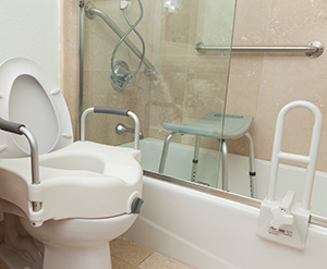 Inodoro con asiento modificado, una silla para la ducha, una barra de sujeción y una regadera de mano en el cuarto de baño.