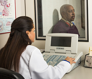 Hombre sentado detrás de una ventana, con auriculares. Proveedora de atención médica del otro lado de la ventana que le realiza una prueba de audición.