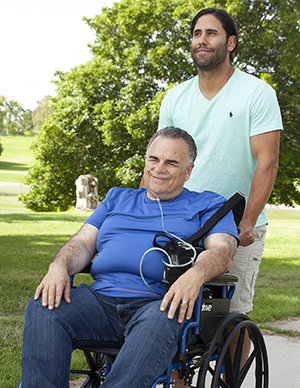 Hombre en silla de ruedas con una cánula nasal. Otro hombre empujando la silla de ruedas.